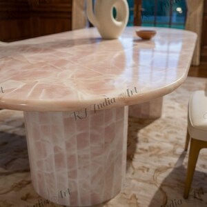 May include: A pink and white oval-shaped dining table with a matching cylindrical base. The table has a glossy finish and a unique pattern. A white vase and a small wooden bowl are on the table. The table is in a well-lit room.