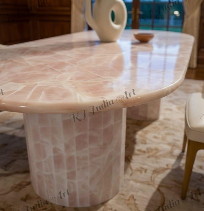 May include: A pink and white oval-shaped dining table with a matching cylindrical base. The table has a glossy finish and a unique pattern. A white vase and a small wooden bowl are on the table. The table is in a well-lit room.