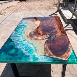 May include: A rectangular wooden table with a unique design. The table features a dark brown wood grain section and a turquoise resin section that resembles an ocean scene. The table is supported by black metal legs.