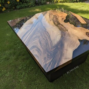 May include: Square coffee table with a unique wood grain design and a glossy, dark epoxy resin finish. The table has a dark base and is set on a green lawn. The wood grain features shades of brown and gray.