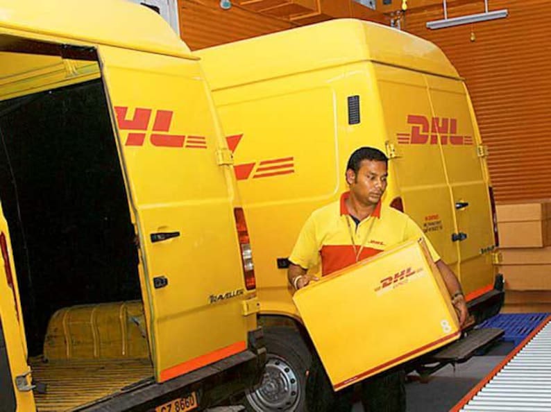May include: A yellow DHL delivery van with the door open, revealing the interior. A person in a yellow and red uniform is holding a yellow DHL box. The DHL logo is visible on the van and the box.