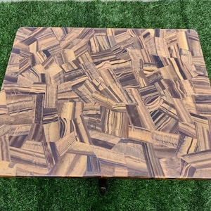 May include: Rectangular coffee table with a mosaic-like surface made of tiger eye stone. The table's surface features a pattern of brown, gold, and black tones. The table is set on a green surface.