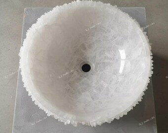 May include: A white, round bathroom sink with a textured, crystalline exterior. The sink has a central drain hole and a rough, jagged edge. The sink is set on a gray surface.