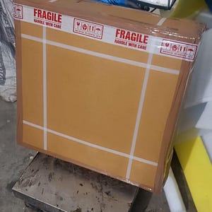 May include: A large, rectangular cardboard box, secured with white tape, sits on a metal scale. The box is labeled "FRAGILE HANDLE WITH CARE" with international shipping symbols. A digital scale and other items are in the background.