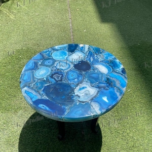 May include: A round, blue agate stone table with a polished surface. The table top is composed of various shades of blue agate, creating a mosaic-like pattern. The table is supported by three black legs. The table measures approximately 76 cm in diameter.
