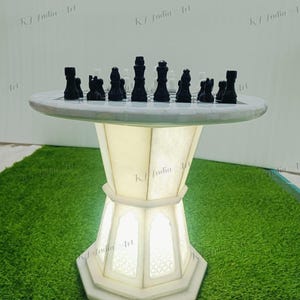 May include: A white marble chess table with black and white chess pieces. The table has a round top and a geometric, illuminated base. The chess pieces are arranged on the board, ready for a game. The table is set on a green surface.