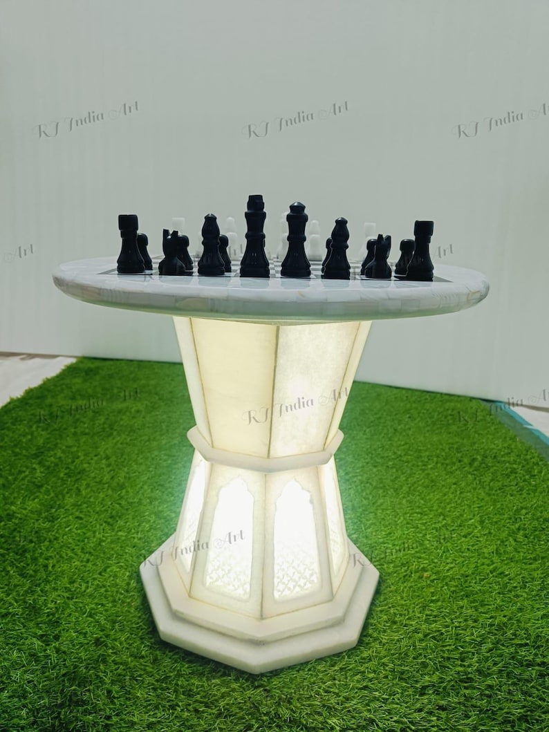 May include: A white marble chess table with black and white chess pieces. The table has a round top and a geometric, illuminated base. The chess pieces are arranged on the board, ready for a game. The table is set on a green surface.