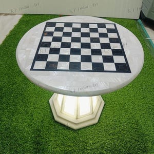 Puede incluir: Una mesa de ajedrez redonda de mármol blanco con una superficie de juego a cuadros blancos y negros. La mesa tiene una base octogonal decorativa. La mesa está sobre una superficie verde.