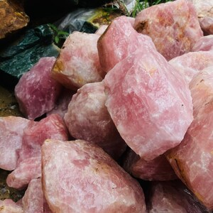 May include: A collection of raw rose quartz crystals in varying sizes and shades of pink. The rough, natural stones have a textured surface and are piled together, showcasing their natural beauty. Some stones have a light pink hue, while others are a deeper rose color.