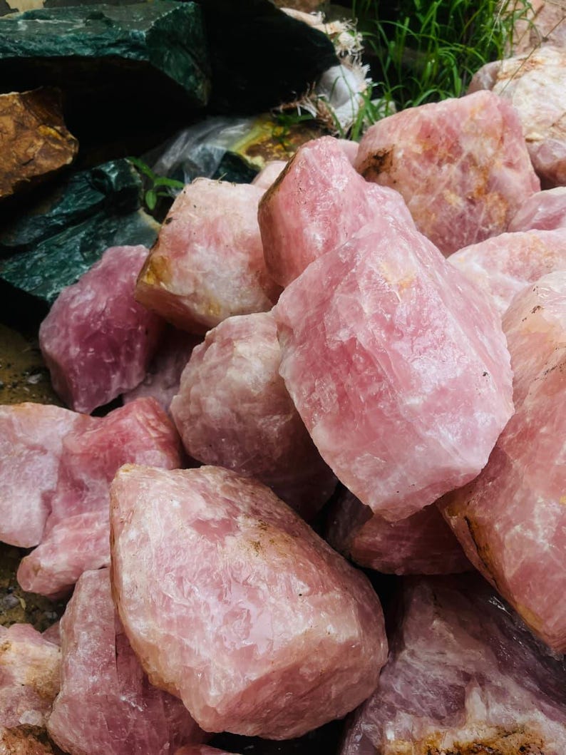 May include: A collection of raw rose quartz crystals in varying sizes and shades of pink. The rough, natural stones have a textured surface and are piled together, showcasing their natural beauty. Some stones have a light pink hue, while others are a deeper rose color.