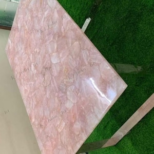 May include: A rectangular rose quartz table with a polished surface. The table is supported by a silver metal frame. The table's surface is a mosaic of pink and white quartz pieces.