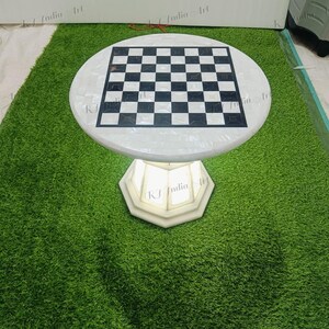 May include: A round, white marble chess table with a black and white checkered playing surface. The table sits on a white, geometric base. The table is displayed on a green, artificial grass surface.