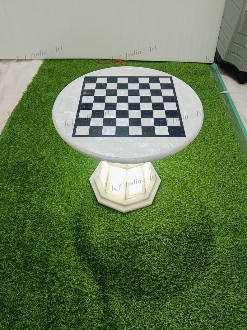 May include: A round, white marble chess table with a black and white checkered playing surface. The table sits on a white, geometric base. The table is displayed on a green, artificial grass surface.