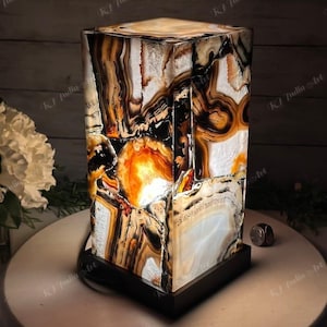 Pode incluir: Uma luminária de mesa retangular com um design único. A luminária apresenta um mosaico de peças de pedra marrom, laranja, preta e branca. Assenta sobre uma base preta e tem um tampo retangular a condizer. A luminária é iluminada por dentro, criando um brilho quente.
