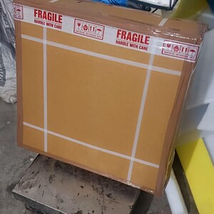 May include: A large, rectangular cardboard box, secured with white tape, sits on a metal scale. The box is labeled "FRAGILE HANDLE WITH CARE" with international shipping symbols. A digital scale and other items are in the background.
