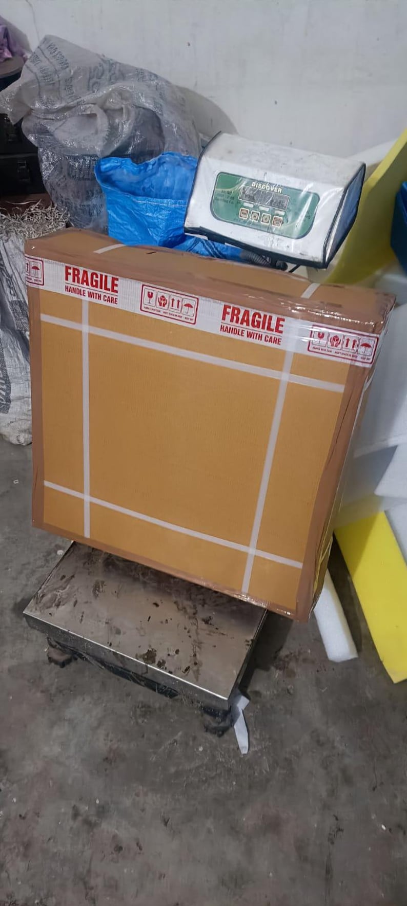 May include: A large, rectangular cardboard box, secured with white tape, sits on a metal scale. The box is labeled "FRAGILE HANDLE WITH CARE" with international shipping symbols. A digital scale and other items are in the background.