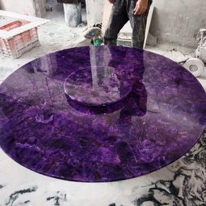 Può includere: Un grande tavolo rotondo con una superficie lucida di colore viola intenso. Il tavolo ha un inserto circolare al centro. La pietra ha un aspetto marmorizzato con venature viola scuro e bianche. Il tavolo si trova in un'officina.