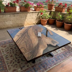 May include: A square wooden coffee table with a dark, glossy finish and a unique wood grain pattern. The table has a clear resin inlay and sits on a black metal base. The table is placed on a patterned rug.