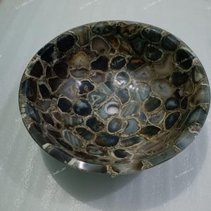 May include: Oval-shaped stone sink with a mosaic pattern. The sink is made of polished, multi-colored stones in shades of gray, brown, and white. The sink has a drain hole in the center and a smooth, rounded rim. A unique bathroom fixture.