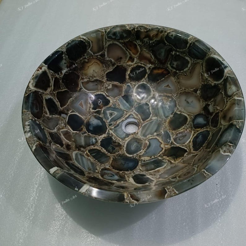 May include: Oval-shaped stone sink with a mosaic pattern. The sink is made of polished, multi-colored stones in shades of gray, brown, and white. The sink has a drain hole in the center and a smooth, rounded rim. A unique bathroom fixture.