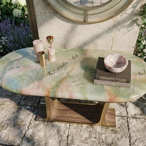 May include: An oval console table with a polished green and pink onyx stone top featuring the text RJ India Art. The base is constructed from curved wood with a metallic finish. On the surface sit two cylindrical candle holders and a small bowl.