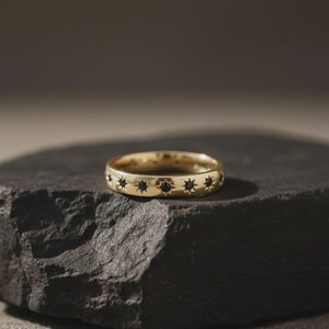 May include: A gold-colored ring with a band featuring a repeating pattern of black star-shaped accents. The ring is displayed on a dark, textured surface, showcasing its design and craftsmanship. The ring's design is simple yet elegant.