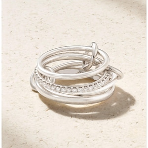 May include: A silver ring featuring multiple bands, some plain and others set with small, clear stones. The bands are interwoven, creating a layered design. The ring is displayed on a light-toned surface.
