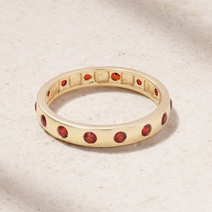 May include: A gold-colored ring with a band of small, round, red gemstones. The ring is set with evenly spaced gemstones around the entire band. The ring is displayed on a light-colored surface.