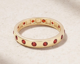 May include: A gold-colored ring with a band of small, round, red gemstones. The ring is set with evenly spaced gemstones around the entire band. The ring is displayed on a light-colored surface.