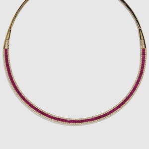 Op de afbeelding: Een gouden ketting met een centrale band van rechthoekige, dieprode edelstenen. De edelstenen worden begrensd door een rij kleine, heldere stenen. De ketting heeft een flexibel ontwerp en een sluiting.