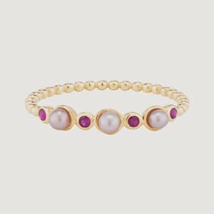 May include: A gold ring featuring a beaded band design. The ring is adorned with three light-colored pearls and four small, round, red gemstones. The ring is set against a light background.