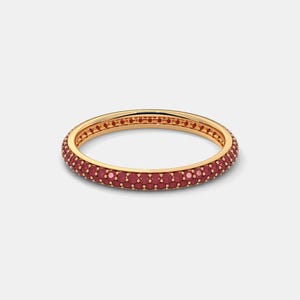 May include: A gold-colored ring with a band of small, round, red gemstones. The ring is set against a plain white background, highlighting the jewelry's design and color. The ring is a piece of jewelry.