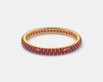 May include: A gold-colored ring with a band of small, round, red gemstones. The ring is set against a plain white background, highlighting the jewelry's design and color. The ring is a piece of jewelry.