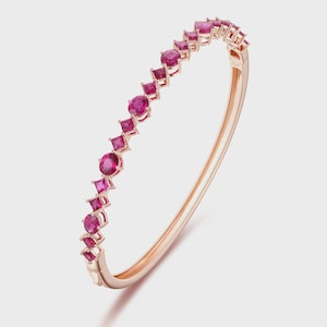 May include: Rose gold bangle bracelet featuring a series of alternating round and square-cut ruby gemstones. The gemstones are set in a linear pattern along the top of the bracelet. The bracelet has a hinged clasp.