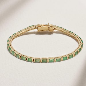 Peut inclure: Un bracelet en or orné de pierres précieuses vertes de forme rectangulaire. Le bracelet est doté d'un fermoir en or et est posé sur une surface blanche. Les pierres sont espacées de manière uniforme.