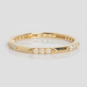 May include: A gold band ring with small, round, white gemstones set around the band. The ring has a polished finish and is set against a white background. The gemstones are evenly spaced around the ring.