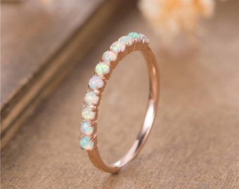 May include: Rose gold ring featuring a row of opal gemstones. The ring's band is a warm rose gold color, and the opals have a milky white base with flashes of green and blue. The ring is set on a neutral background.