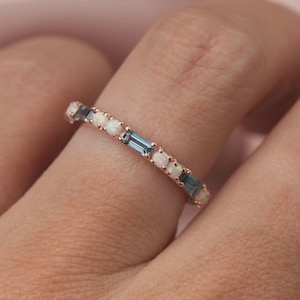 May include: A rose gold ring featuring alternating white opal and rectangular blue gemstones. The ring is set with multiple small stones, creating a delicate and elegant design. The ring is shown on a finger.