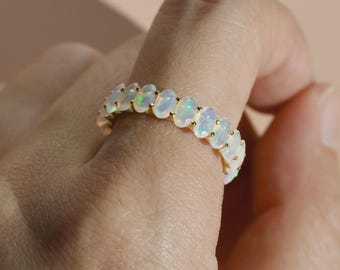 May include: A gold ring featuring a series of oval-shaped opals. The opals are white with iridescent flashes of color. The ring is set on a gold band, creating a delicate and elegant design.