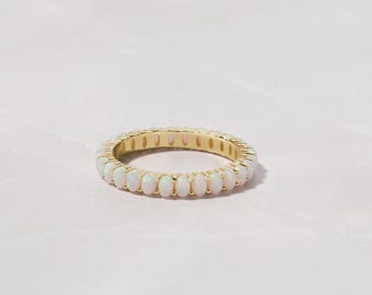 May include: A gold ring featuring a continuous band of oval-shaped, white opal gemstones. The ring's gold band provides a warm contrast to the opals, creating a classic and elegant look. The ring is a piece of jewelry.