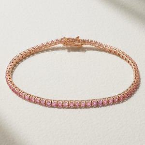 Könnte beinhalten: Ein roségoldenes Tennisarmband mit einer durchgehenden Reihe rosafarbener Edelsteine. Das Armband hat einen sicheren Verschluss. Die Edelsteine sind gleichmäßig angeordnet und verleihen Glanz.