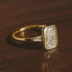 May include: A gold ring with a large, rectangular, clear gemstone as the primary focus. A smaller, rectangular gemstone is set on either side of the main stone. The ring is set on a dark wooden surface.