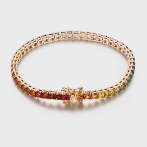 Könnte beinhalten: Goldfarbenes Armband mit einer Reihe von bunten, quadratisch geschliffenen Edelsteinen. Die Edelsteine wechseln durch ein Farbspektrum. Das Armband hat einen sicheren Verschluss. Ca. 18 cm lang.