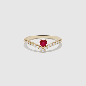 May include: A gold V-shaped ring featuring a central heart-shaped red gemstone flanked by a row of small, clear round-cut stones along the band. The design creates a chevron silhouette with a polished metal finish and prong settings.