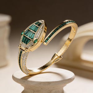 Könnte beinhalten: Ein goldfarbenes Armband in Schlangenform mit smaragdgrünen und klaren Kristallakzenten. Der Schlangenkopf ist mit grünen Steinen besetzt, und das Armband ist mit Kristallen verziert. Das offene Armband liegt auf einem kleinen, cremefarbenen Sockel.