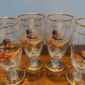 May include: Four clear wine glasses with gold rims and pheasant designs. Each glass has a flying pheasant and a perched pheasant in brown, orange, and yellow. The glasses have a classic stem and base, suitable for a dinner party.