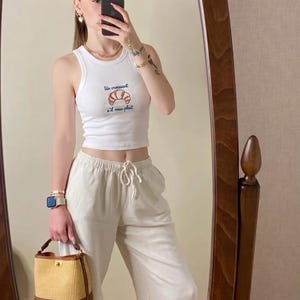 Pode incluir: Top curto branco com um gráfico de croissant e o texto "Un croissant s'il vous plaît". O top é combinado com calças de linho bege e uma pequena bolsa de palha com acabamento em couro castanho.