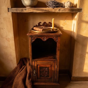 May include: A dark wood cabinet with a single door and open shelf, topped with a brass tray holding a leather-bound book and a yellow candle. A brown herringbone throw blanket rests beside the cabinet, with a bowl and dried lavender above.