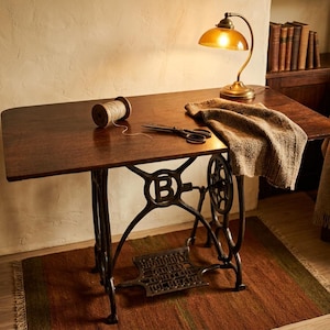 May include: A dark brown wooden sewing machine table with a black cast iron treadle base featuring a prominent letter B. On the tabletop sits a wooden spool of thread, black metal scissors, and a folded beige knit fabric under a brass desk lamp.