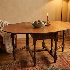 May include: A dark brown wooden drop-leaf table with an oval top and turned legs. The table features a central rectangular base with a drawer and hinged side leaves. A green bowl with three round decorative spheres sits on the table surface.